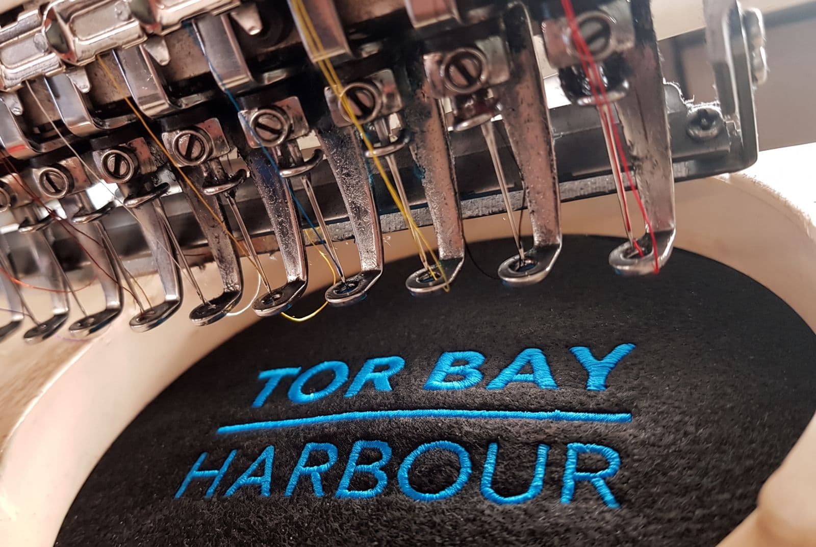 Embroidery machine stitching the Tor Bay Harbour logo in blue thread onto black fleece